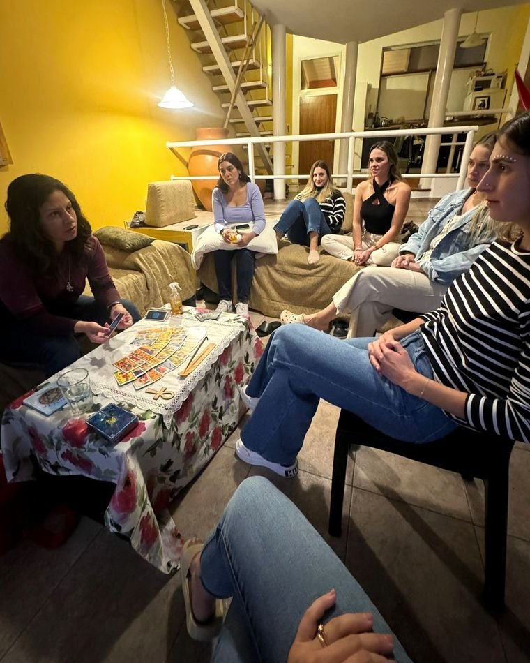 Mariana Medici sentada frente a una mesa baja con la tirada de Tarot de Marsella abierta, mirando a seis mujeres sentadas en ronda en el living de la casa