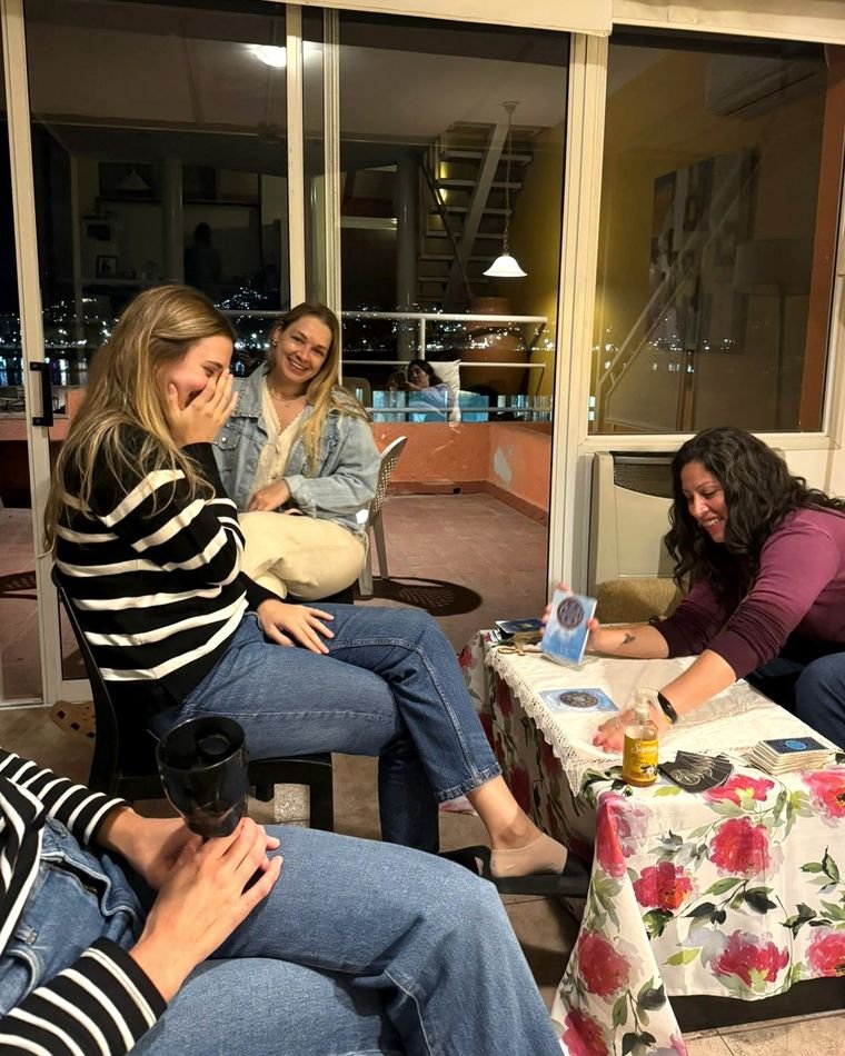 Mariana Medici mostrando una carta a dos amigas que se ríen, junto al ventanal de una terraza con las luces de la ciudad de fondo